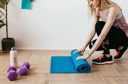 Side view of young female athlete in sportswear unfolding blue sport mat and looking down while sitting on floor near dumbbells and water bottle preparing for training indoors